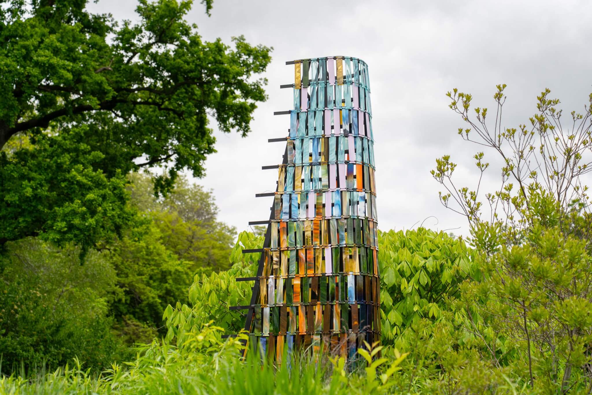 A tapering tower made of colored glass panels rises from a meadow at Brooklyn Botanic Garden.