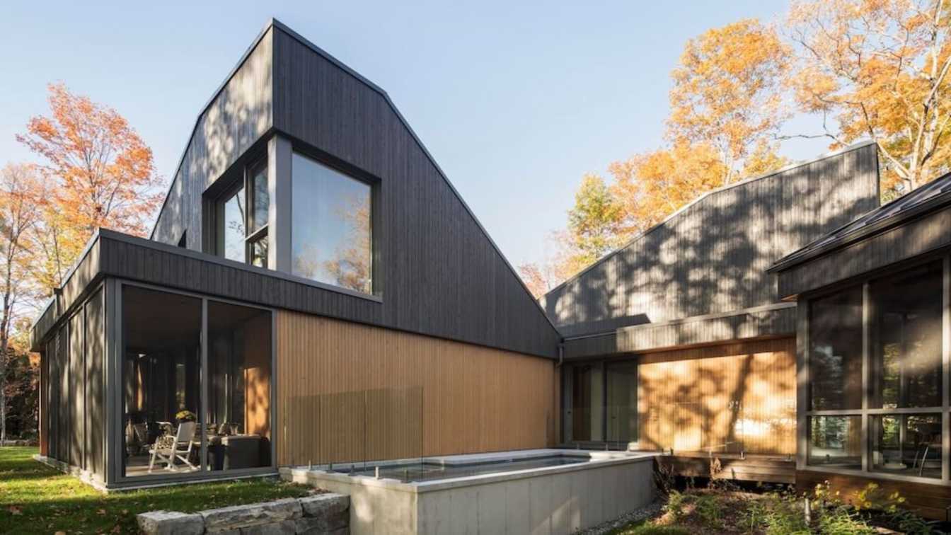 Exterior view of L'Échouage Residence by Bourgeois / Lechasseur architectes, showing the original cottage and new pavilions clad in two-toned cedar, nestled by the St. Lawrence