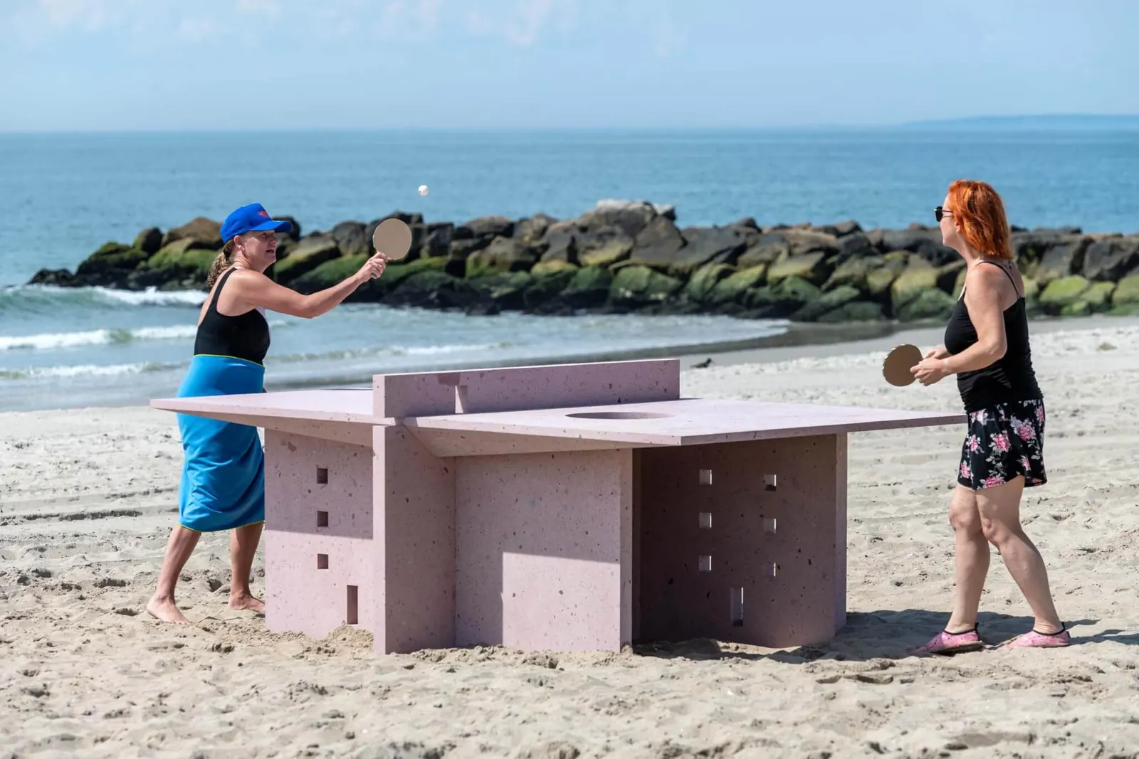 An outdoor art exhibition on Rockaway Beach, featuring several uniquely designed ping-pong tables, with some tables incorporating sculptural elements and others featuring colorful