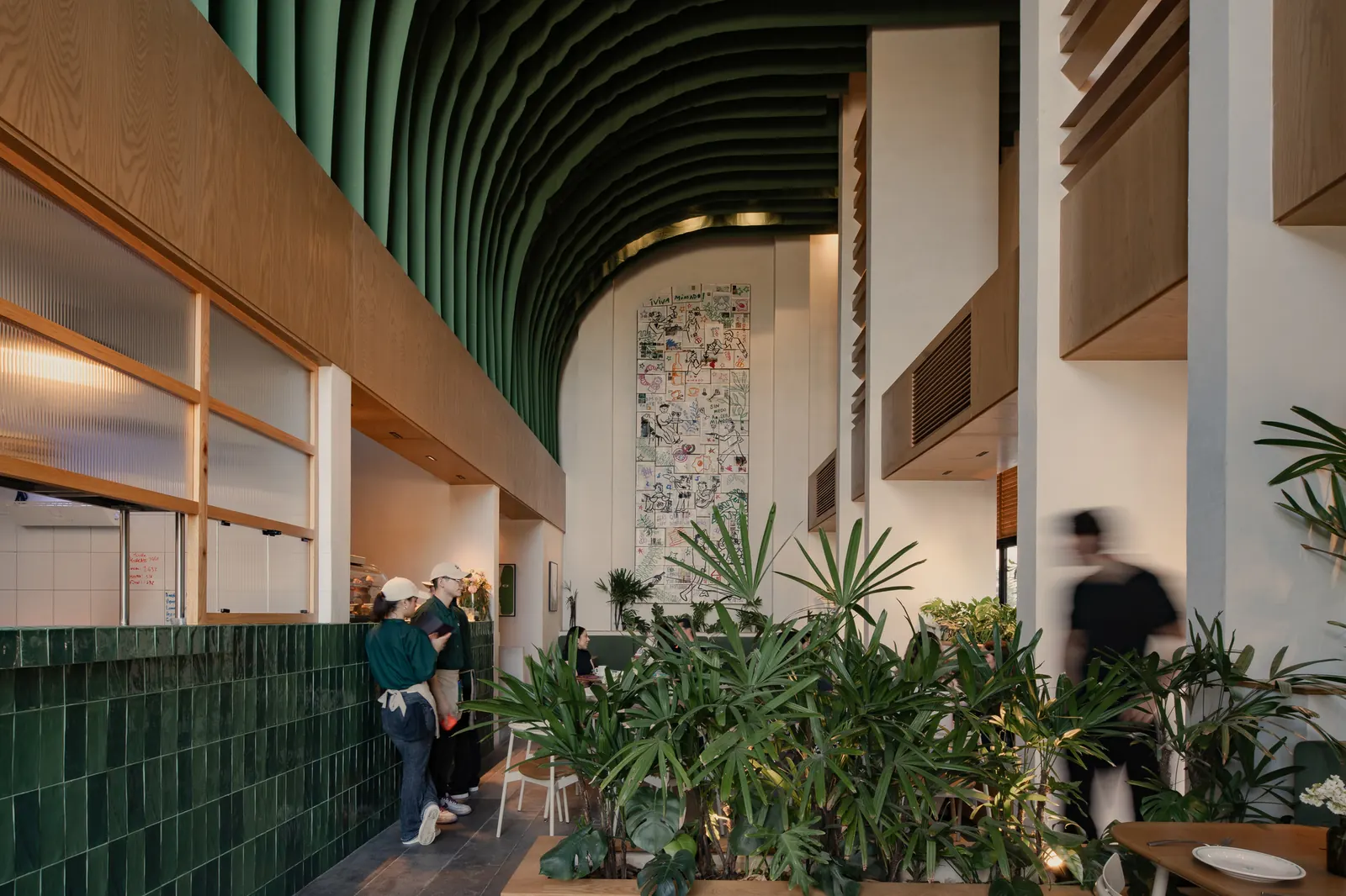 Interior view of Mimado restaurant in Mérida showing green ceramic tile walls, white oak furniture, fluted glass partitions, and suspended fabric panels under a double-height