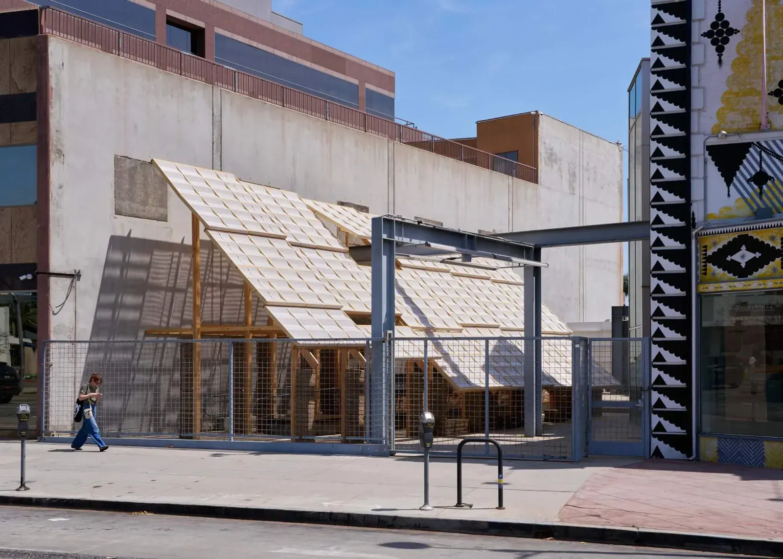 A view of the "Earthen Comforts: Airing Earths" installation, showcasing its wooden structure, earthen block supports, and woven canopy designed for shade and airflow.