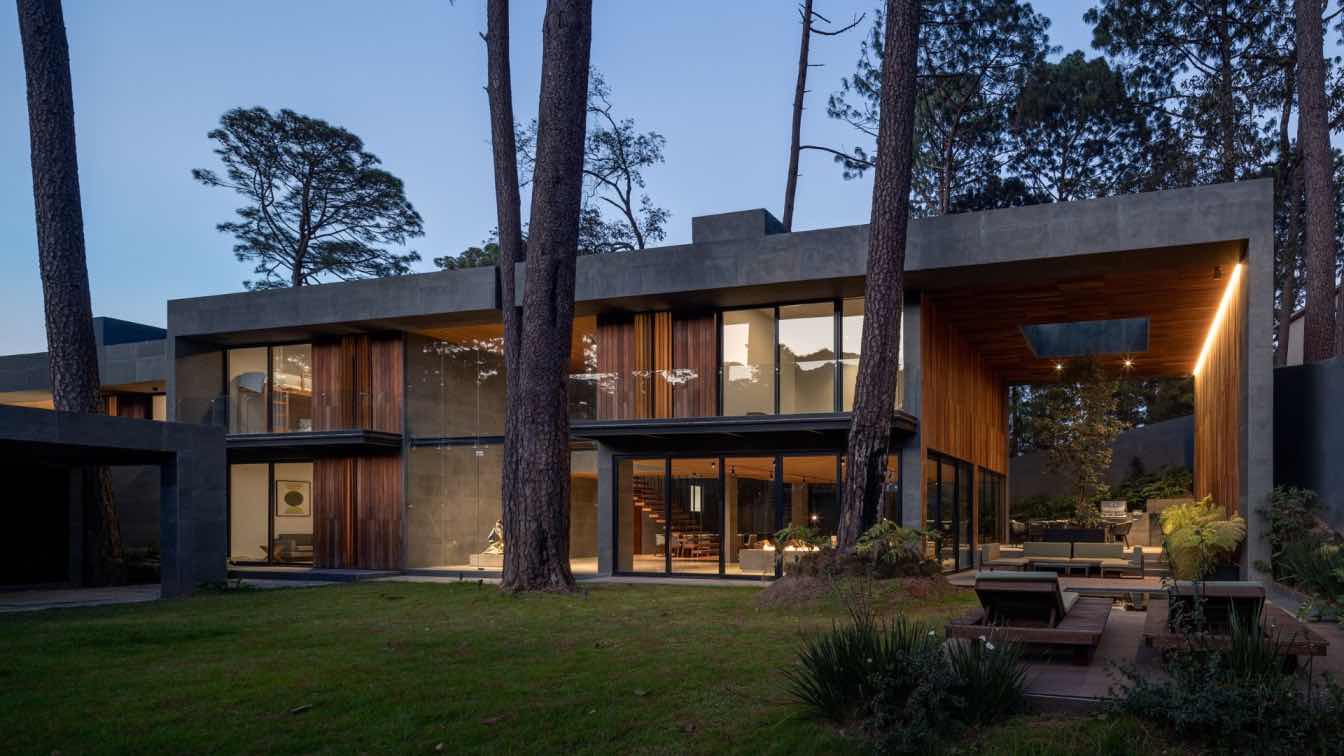 Exterior view of Casa Valle, a modern retreat in Valle de Bravo, Mexico, with large stone frames and expansive glass windows, surrounded by lush forest.